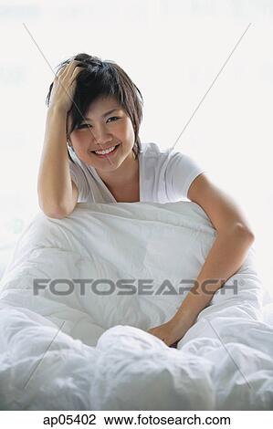 Stock Image - Woman sitting on bed, smiling at camera. Fotosearch