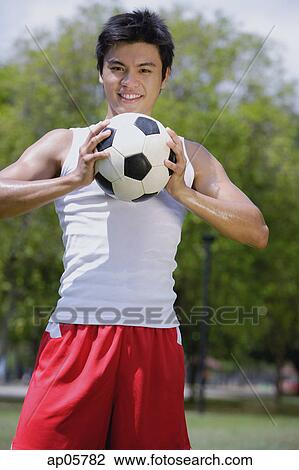 Stock Image - Young man holding soccer ball, looking at camera. Fotosearch