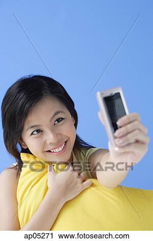 Stock Image - Young woman hugging pillow, using camera phone. Fotosearch