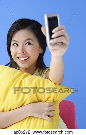 Stock Image - Young woman hugging pillow, using camera phone. Fotosearch