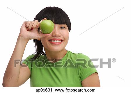 Stock Image - Young woman in green T-shirt holding green apple over eye. Fotosearch