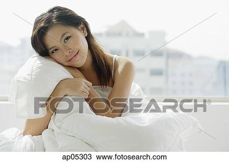 Stock Image - Young woman sitting on bed, embracing pillow. Fotosearch