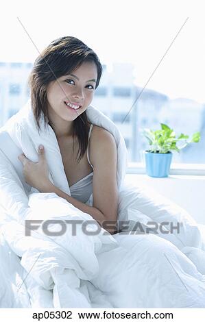 Young woman sitting on bed, wrapped in a blanket, smiling at camera View Large Photo Image Stock Image - Young woman sitting on bed, wrapped in a blanket, smiling at camera. Fotosearch