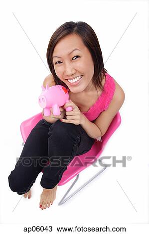 Stock Image - Young woman sitting on chair, holding piggy bank, smiling. Fotosearch
