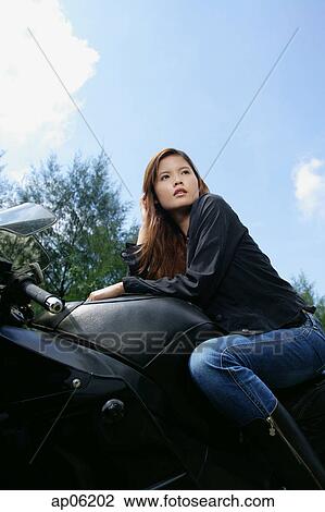 Stock Image - Young woman sitting on motorcycle, looking away, low angle view. Fotosearch