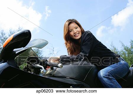 Stock Image - Young woman sitting on motorcycle, smiling at camera, low angle view. Fotosearch