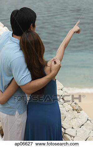Stock Photo - Couple looking out at sea, rear view, woman pointing in the distance. Fotosearch