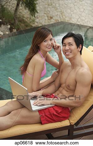 Stock Photo - Couple sitting by swimming pool, man using laptop, woman on the phone, both smiling at camera. Fotosearch