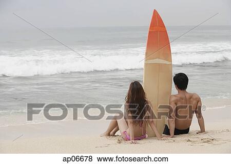 Stock Photo - Couple sitting on beach, surfboard between them, looking out to sea. Fotosearch