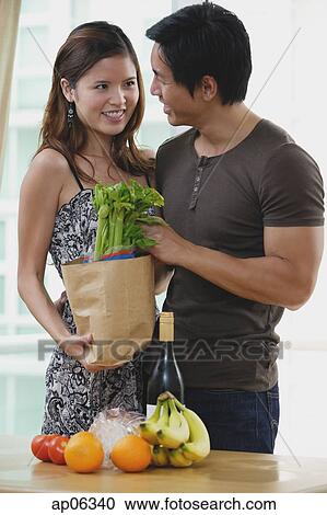 Stock Image - Couple smiling at each other, woman holding shopping bag with groceries. Fotosearch