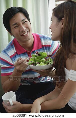 Stock Image - Couple talking, man eating a salad, woman holding a mug. Fotosearch