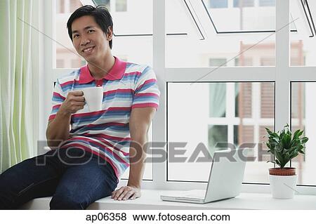 Stock Photo - Man sitting on bay window, holding a cup, laptop open next to him. Fotosearch