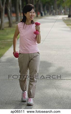 Stock Image - Mature woman using dumbbells, walking along path. Fotosearch