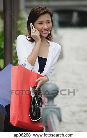 Stock Photo - Woman leaning on railing, carrying shopping bags, using mobile phone. Fotosearch