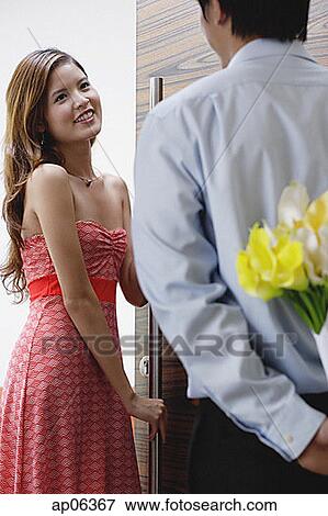 Stock Photo - Woman opening door for man, man hiding flowers behind his back. Fotosearch