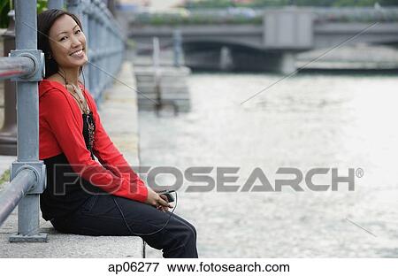 Woman sitting by river, listening to MP3 player, smiling at camera View Large Photo Image Stock Photo - Woman sitting by river, listening to MP3 player, smiling at camera. Fotosearch
