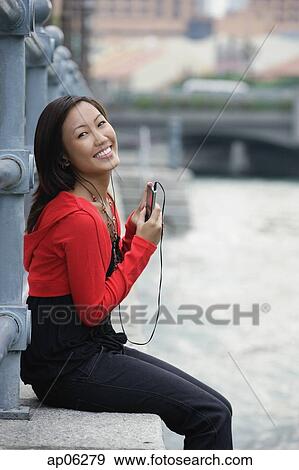 Stock Photo - Woman sitting by river, listening to MP3 player, portrait. Fotosearch