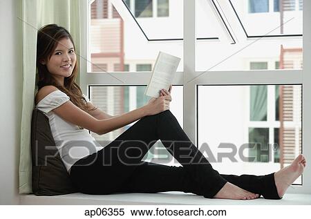 Woman sitting by window holding a book, smiling at camera View Large Photo Image Stock Photography - Woman sitting by window holding a book, smiling at camera. Fotosearch