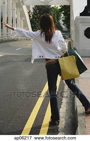Stock Photo - Woman standing by road, waving for taxi, rear view. Fotosearch