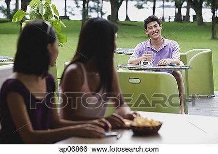 Young man sitting at outdoor cafe, smiling at women in the foreground View Large Photo Image Stock Photograph - Young man sitting at outdoor cafe, smiling at women in the foreground. Fotosearch