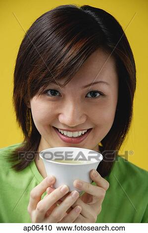 Stock Image - Young woman holding Chinese tea cup, smiling at camera. Fotosearch