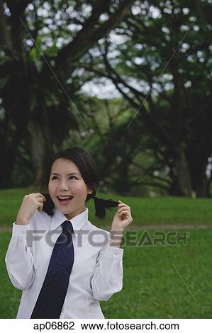 Stock Image - Young woman in school uniform, standing in park, touching her hair. Fotosearch