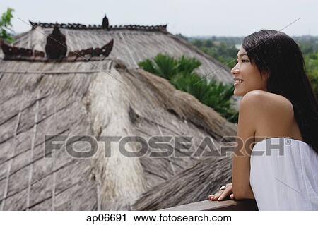 Stock Image - Young woman leaning on railing looking out at thatched roofs. Fotosearch