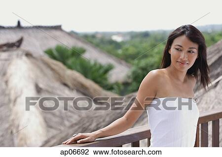Stock Image - Young woman leaning on railing looking away, thatched roofs behind her. Fotosearch