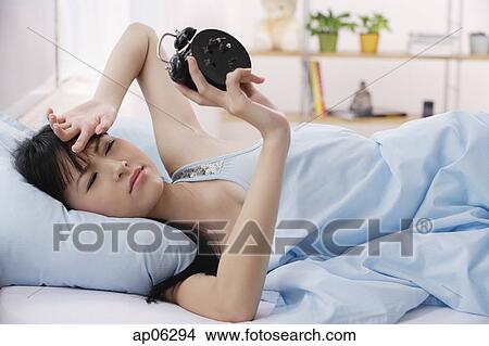 Picture - Young woman lying on bed, frowning at alarm clock. Fotosearch