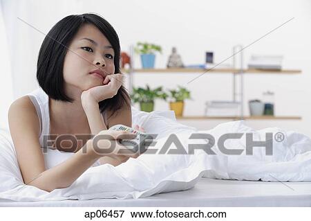 Young woman lying on bed, hand on chin, holding TV remote control View Large Photo Image Stock Photo - Young woman lying on bed, hand on chin, holding TV remote control. Fotosearch
