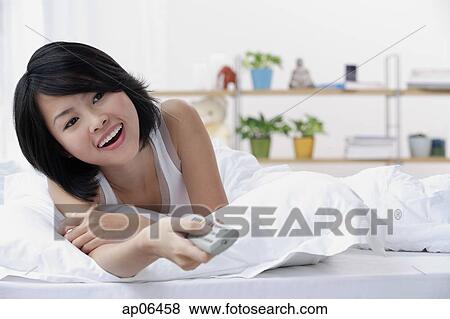 Stock Photo - Young woman lying on bed, holding TV remote control. Fotosearch