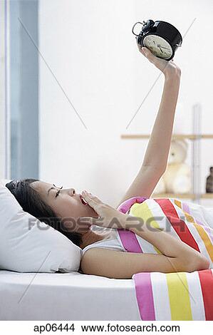 Picture - Young woman lying on bed, looking at alarm clock, hand on mouth. Fotosearch