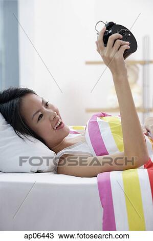 Stock Image - Young woman lying on bed, looking at alarm clock, smiling. Fotosearch