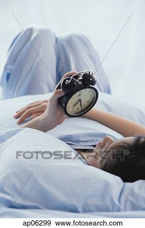 Stock Photo - Young woman lying on bed, looking at alarm clock. Fotosearch