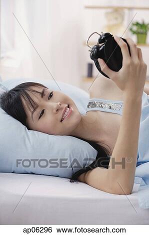 Young woman lying on bed, looking at alarm clock, smiling View Large Photo Image Stock Photograph - Young woman lying on bed, looking at alarm clock, smiling. Fotosearch