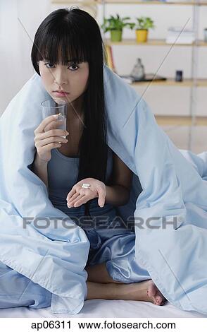 Stock Image - Young woman on bed, holding glass of water and pills. Fotosearch