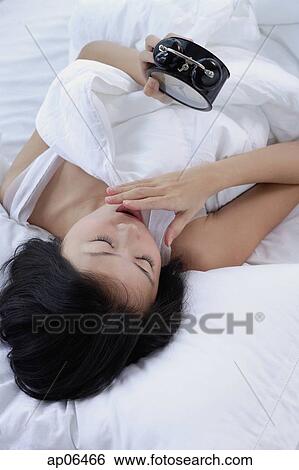 Young woman on bed, looking at alarm clock, hand on mouth View Large Photo Image Stock Photograph - Young woman on bed, looking at alarm clock, hand on mouth. Fotosearch