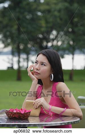 Stock Image - Young woman sitting at outdoor table, hand on chin, looking away. Fotosearch