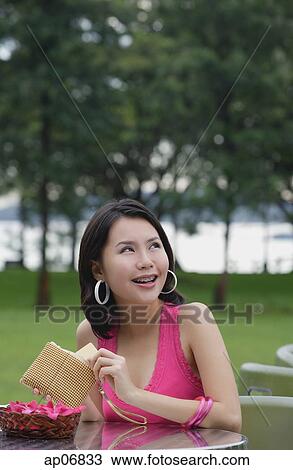 Stock Image - Young woman sitting at outdoor table, holding wallet, looking away. Fotosearch