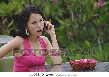 Young woman sitting at table outdoors, on the phone, angry expression on face View Large Photo Image Stock Image - Young woman sitting at table outdoors, on the phone, angry expression on face. Fotosearch