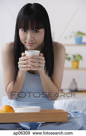Young woman sitting on bed, holding cup of coffee View Large Photo Image Stock Photo - Young woman sitting on bed, holding cup of coffee. Fotosearch