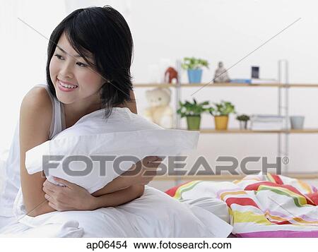 Picture - Young woman sitting on bed, hugging pillow, looking away. Fotosearch
