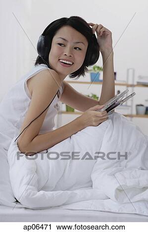 Stock Image - Young woman sitting on bed, listening to music. Fotosearch