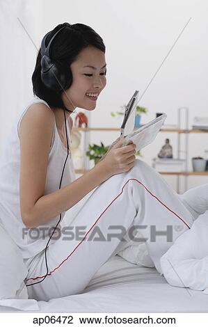 Young woman sitting on bed, listening to music, smiling View Large Photo Image Stock Image - Young woman sitting on bed, listening to music, smiling. Fotosearch