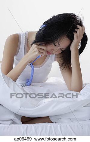 Young woman sitting on bed, using telephone, hand on head View Large Photo Image Stock Photo - Young woman sitting on bed, using telephone, hand on head. Fotosearch