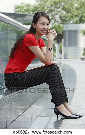 Stock Photo - Young woman sitting on ledge outdoors, smiling at camera. Fotosearch