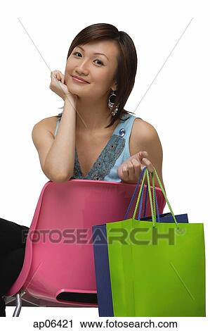 Stock Image - Young woman sitting on pink chair, holding shopping bags, smiling at camera. Fotosearch