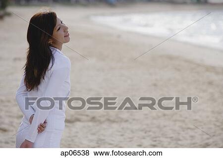 Young woman standing on beach, hands behind back, looking away View Large Photo Image Stock Photo - Young woman standing on beach, hands behind back, looking away. Fotosearch