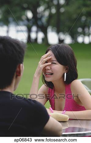 Stock Image - Young woman with hand over face, smiling at man sitting opposite from her. Fotosearch