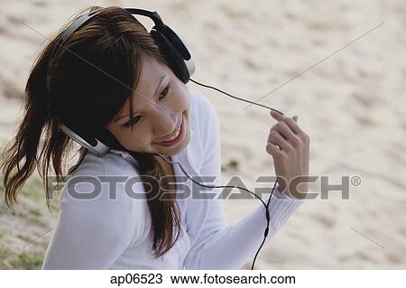 Stock Image - Young woman with head phones, sitting on beach. Fotosearch
